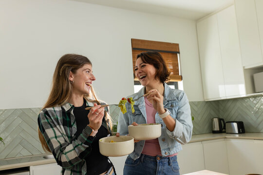Diverse female friends laughing and sharing salad with forks from ceramic bowls in home kitchen