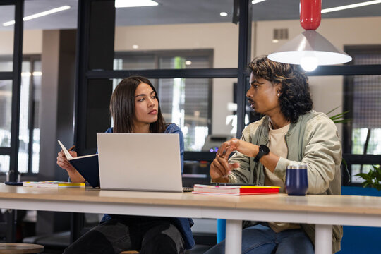 Asian coworkers sitting at shared table in modern coworking office, reviewing laptop and notebooks