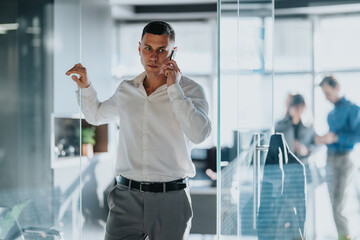 A business professional speaks on his mobile inside a glass-walled office. Colleagues work in the...