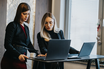 Two professional women collaborate at a standing desk with laptops in a modern office. They review information on screen and discuss at the workstation.