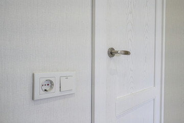 A white interior door with a sleek metallic handle, adjacent to a white wall socket with a power outlet and light switch. Clean, minimalist design.