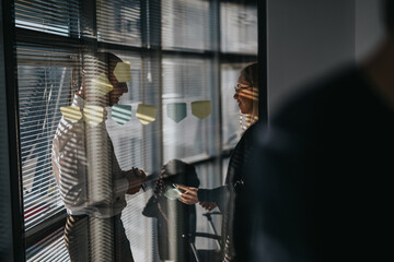 Two professionals converse by a glass wall in a modern office, surrounded by sticky notes and reflections.