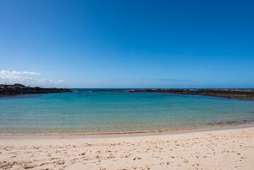 Obraz premium Pale sand meets aquamarine water and dark basaltic outcrops in Lanzarote. Soft waves and footprints mark a quiet moment under a clear sky in wide horizontal light.