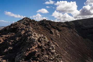 Obraz premium A rugged volcanic ridgeline forms a crater crest in Timanfaya, Lanzarote, Canary Islands. Layered lava strata show in daylight with deep shadows and no vegetation.