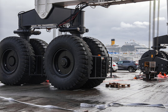 Ground level view shows multi axle wheels, cables, and hydraulics of a mobile boat hoist on a wet apron. A white cruise ship with a yellow funnel docks in Las Palmas.