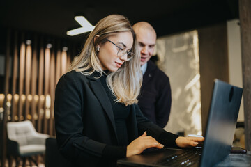 A businesswoman types on a laptop while a coworker observes in a professional office. The scene conveys focus, teamwork, and a contemporary work environment.