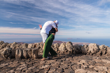 A couple embraces on a cobblestone lookout at Mirador del Rio, Lanzarote. Rugged volcanic rock frames the north coast and Chinijo islets under warm afternoon light.
