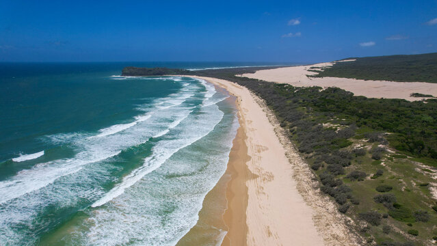 Aerial view of waves crashing onto the sandy shore next to the lush green vegetation with Tukkee Sandblow and Indian Head in the background, K'Gari Fraser Island, Queensland, Australia.