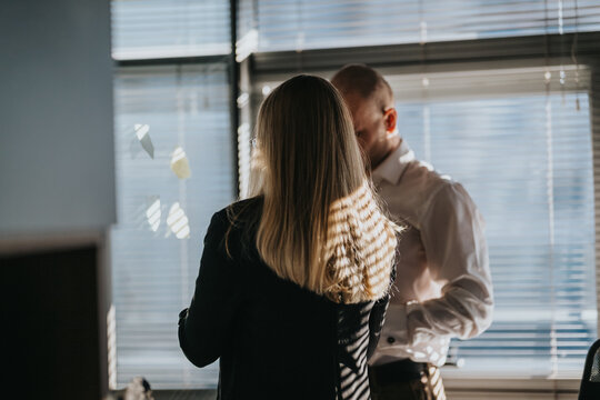 Two people in an office share a conversation near a window as light casts striped shadows. A female with long hair faces a male colleague in a white shirt, creating a candid, collaborative moment.