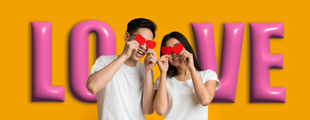 A young couple smiles while holding red heart shapes in front of their faces. Bright letters spell out love in the background. They appear joyful in a playful setting.