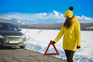 Woman standing by a broken car on a snowy road, placing a red warning triangle
