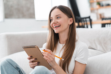 Pretty young Caucasian woman sit in bedroom on floor holds pen thinks over future business plan, take notes in paper notebook, write to-do list creative ideas looking inspired, study concept