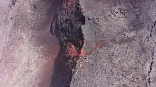 Drone footage ascending over Dolomieu Crater at Piton de la Fournaise on Reunion Island, revealing a wide view of fractured volcanic rock formations.Fog drifts and tiny hikers are visible.