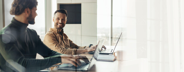 Two men sit at a desk with laptops working on tasks. They are smiling and engaged in their work....