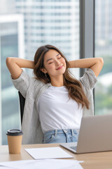 Beautiful Woman relaxing in an office chair with her hands behind her head, laptop and coffee cup on the desk in a bright office setting