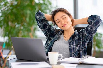 Beautiful Woman relaxing in an office chair with her hands behind her head, laptop and coffee cup on the desk in a bright office setting