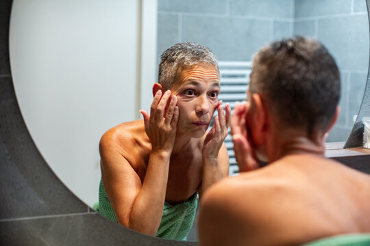Mature woman examining face in home bathroom mirror
