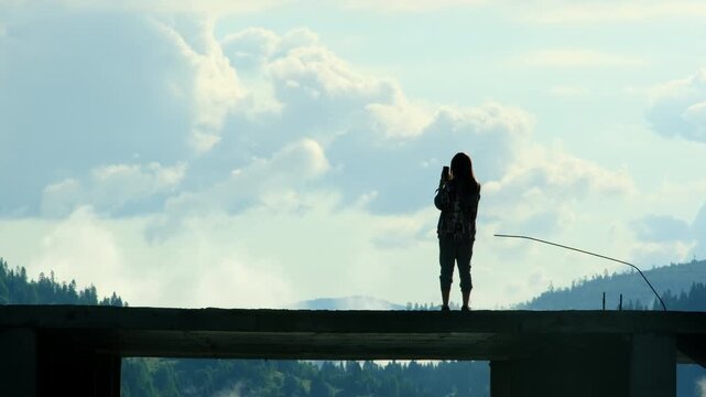 Silhouette of woman taking picture on smartphone on abandoned building top. Lady contemplates mountain scene from construction site slow motion