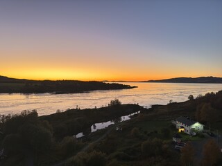 Atmospheric aerial view of green forest with trees and small house at dusk. Beyond it, wide fjord and sky with bright orange sunset. Nature, calm atmosphere, scenic landscape, travel, adventure.
