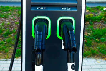 Two charging ports at a station for electric vehicles stand in a parking lot. The green lights indicate readiness for use. The area includes grass and paved surfaces.
