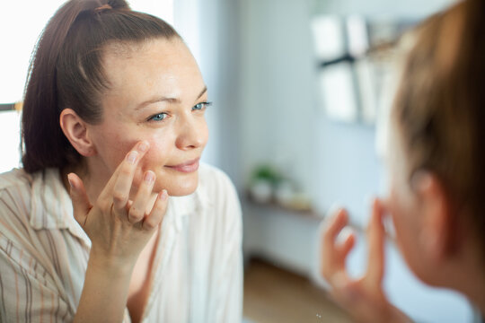 Young woman applying face cream at home in front of mirror