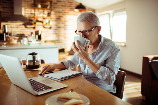 Older woman drinking coffee while working on laptop in home kitchen