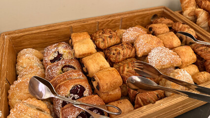 Assorted fresh pastries and croissants with powdered sugar on wooden tray buffet display with serving tongs for breakfast or brunch