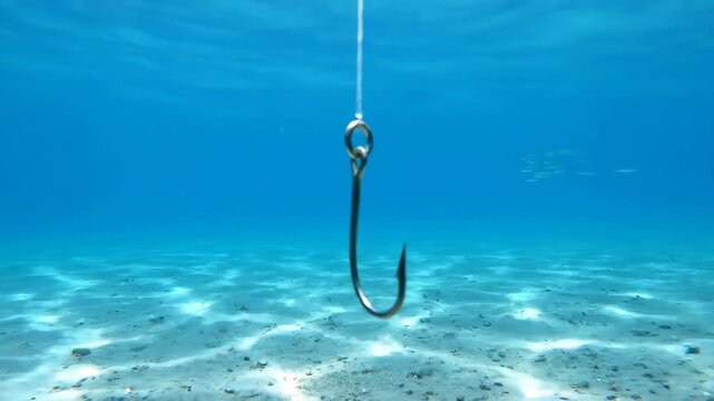 Fishing Hook Underwater - A silver fishing hook hangs suspended by a clear string in clear blue ocean water. The sandy bottom of the sea is visible below.
