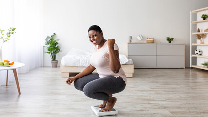 Excited curvy black woman sitting on scales at home, making YES gesture, happy with result of her...