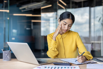 Tired young mature Indian businesswoman sitting at a desk in the office and working with documents