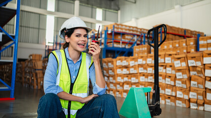 Professional female warehouse worker using radio communication and sitting on pallet jack in a storage facility. Team of logistics experts managing inventory and industrial supply chain.