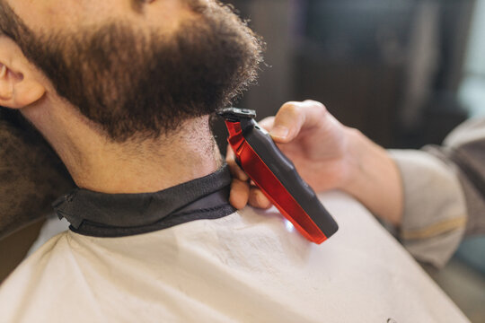Barber trimming beard of male client with electric clipper in barbershop. Professional grooming process, beard shaping, men haircare routine, classic barber service.