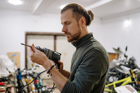 A man inspects a drill in his workshop, where he is surrounded by various bicycles and an array of tools as he works