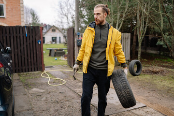 Man changing a car tire in a quiet yard on a cloudy day, displaying determination and practical skill © yaaaaan