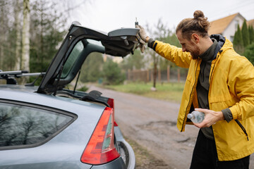 Man opens car trunk on a cloudy day while holding a water bottle near a dirt road surrounded by trees © yaaaaan