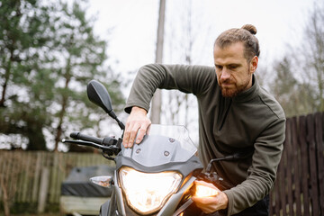 Man works on motorcycle in outdoor setting during overcast day, focusing on light and adjustments in a wooded area