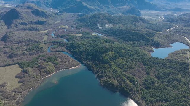 Aerial drone view showing a deep blue lake connected to winding rivers that flow through a dense green forest and mountainous terrain in Patagonia