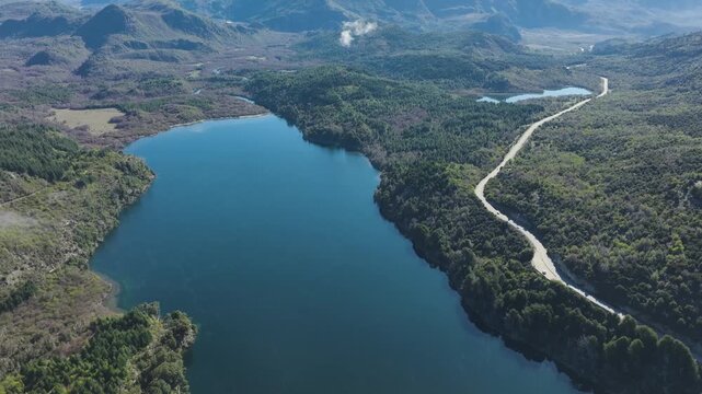 High-altitude aerial drone view of deep blue expanse of Lake Machonico, bordered by dense green forests and a winding section of the Seven Lakes Road in Patagonia, Argentina