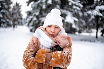 Fototapeta premium Emotional woman looks surprised on snowy path in winter forest. Middle aged female in brown sheepskin coat and white hat. Travel, nature, weekend, lifestyle concept
