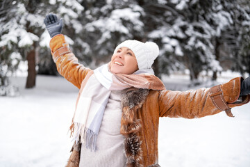 Joyful woman raises hands and looks up in snowy winter forest. Happiness and freedom. Middle aged female in brown coat and white hat. Nature, wellness, lifestyle concept