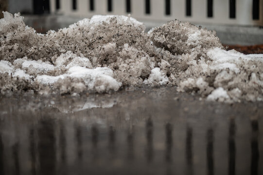 Closeup of dirty, icy snow and slush along the edge of a parking lot with black fence pickets in the background and reflected in a puddle