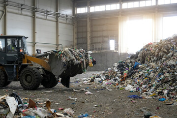Front loader tractor moves a scoop of mixed garbage towards a massive pile in an industrial warehouse. Waste processing, recycling, and pollution concept. Gritty style with bright backlighting
