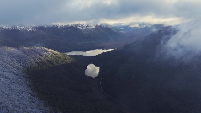 Aerial view looking through clouds at two distinct lakes nestled in deep, forested mountain valleys with snow-dusted slopes in the foreground