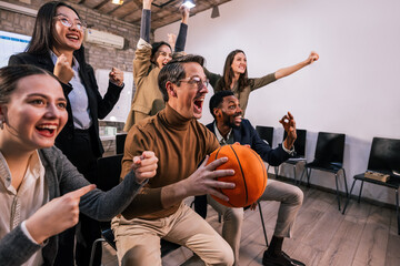 Business team watching a basketball game together at the office, cheering with excitement while one coworker holds a basketball, representing teamwork, relaxation, and positive work atmosphere.