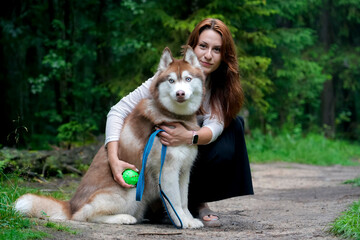 A woman pets and hugs her brown-and-white husky. They are sitting on a dirt path in a lush green forest.