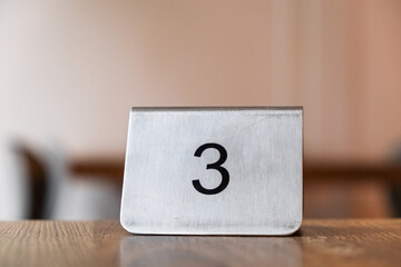 A metal table number '3' sits on a wooden surface, likely in a restaurant or cafe, indicating a table assignment for patrons.
