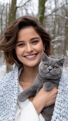 A young woman in a sweater smiles while holding a grey cat against a winter landscape filled with snow and trees