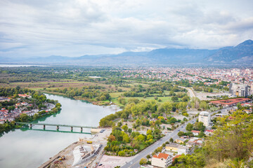 View of an Albanian city surrounded by green riverbanks, a flowing river with a bridge, urban neighborhoods, and distant mountain ranges under a cloudy sky. Scenic cityscape combining nature and urban