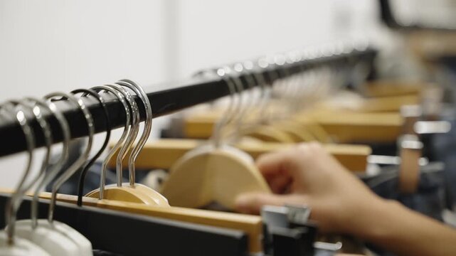 Close-up of a clothing rack lined with wooden and plastic hangers holding denim and assorted garments, in shallow focus as a hand browses organized apparel in a minimalist shop