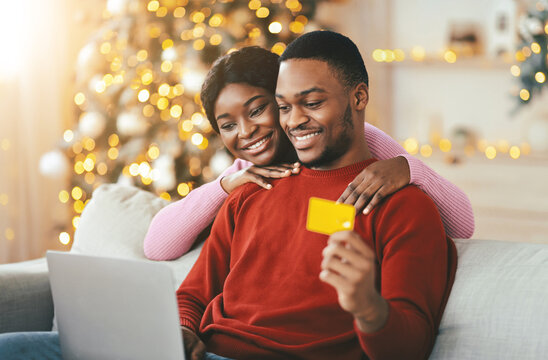 A man and woman smile while sitting on a couch with a laptop during the holiday season. The woman hugs the man from behind as they look at the screen. - Powered by Adobe